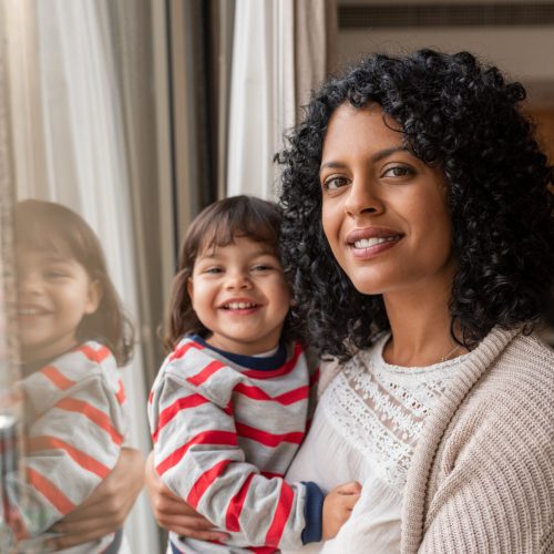 Portrait of a smiling mother holding her cute little daughter in her arms while standing by a window together at home